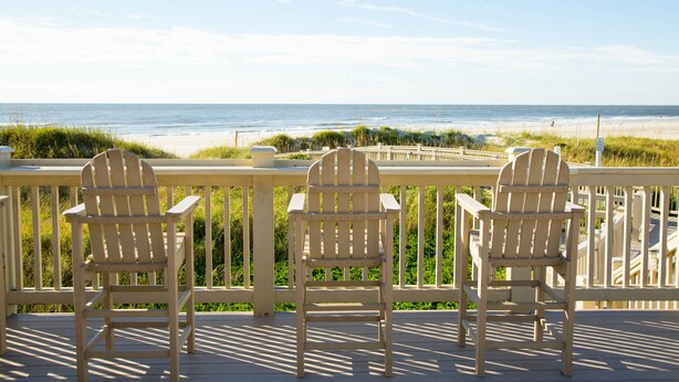 A wooden deck with three Adirondack chairs facing a beach.