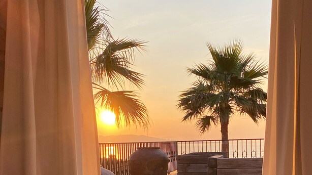 A hotel room with a view of a sunset over a beach, featuring palm trees and a wooden deck. - French stonehouse with fantastic views (Théoule-sur-Mer)