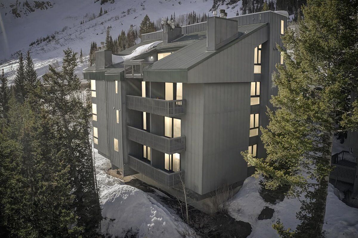 A modern multi-story building with balconies, surrounded by snow-covered mountains and trees.
