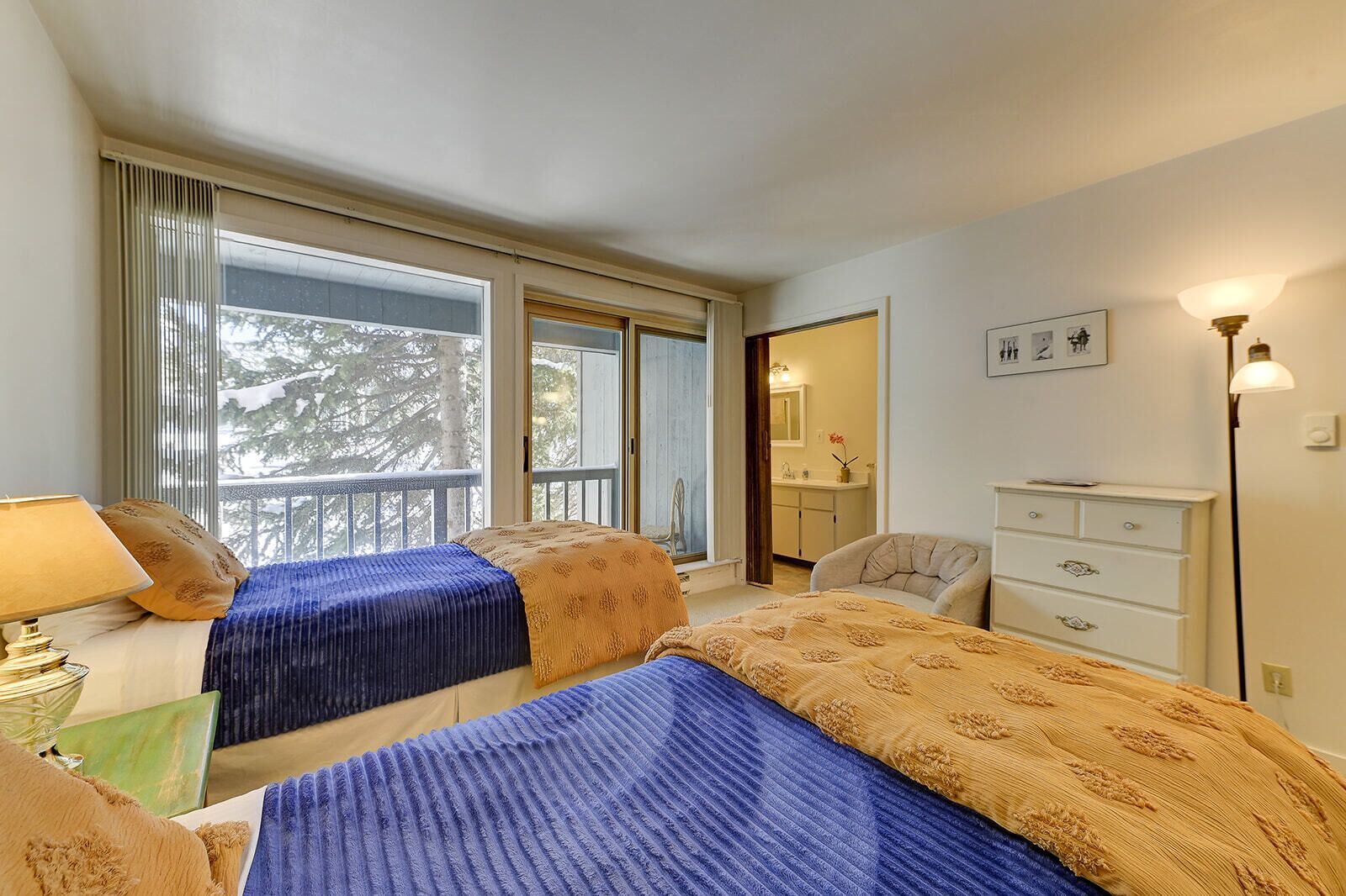 A hotel room with two beds, a dresser, a chair, a lamp, and a window with a view of trees.