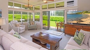 Un salon spacieux doté d’un grand téléviseur, de sièges confortables et offrant une vue sur un jardin luxuriant grâce à de grandes fenêtres.