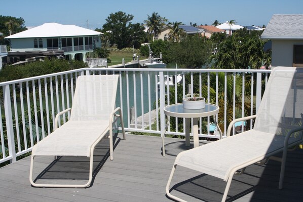 A deck with two white lounge chairs and a small round table, overlooking a body of water and a residential area with houses and palm trees.
