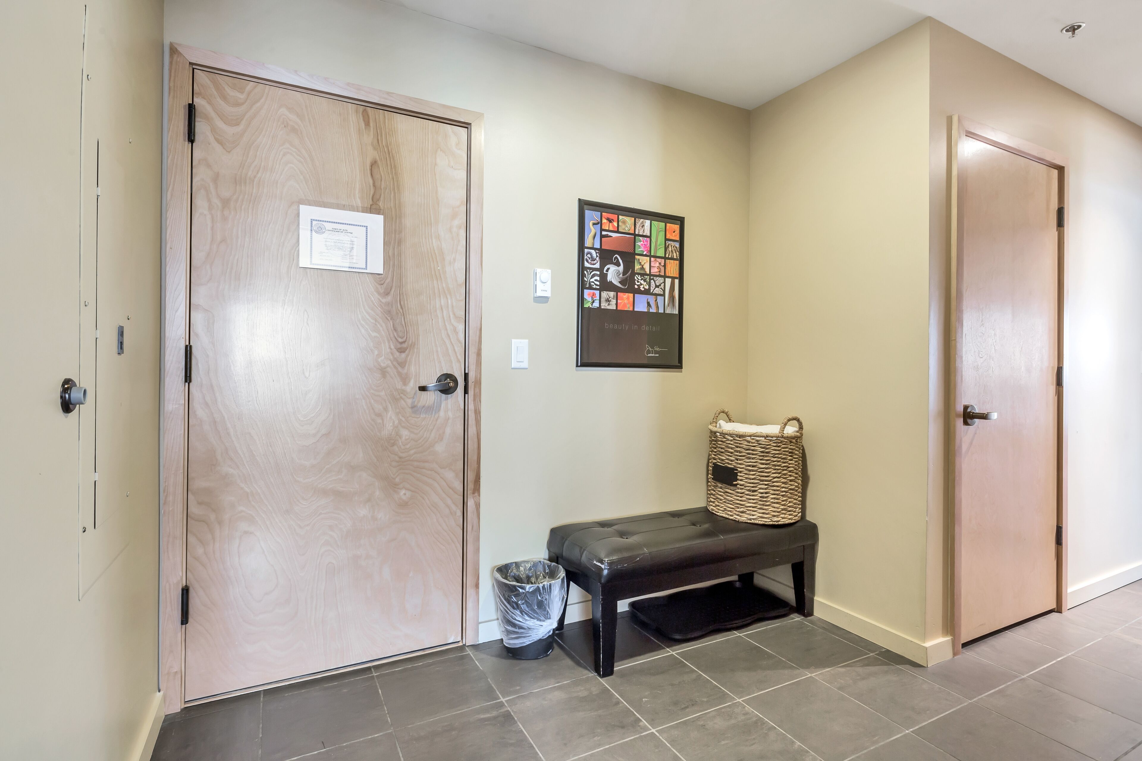 A hallway with two wooden doors, a bench, a trash can, and a framed artwork on the wall.