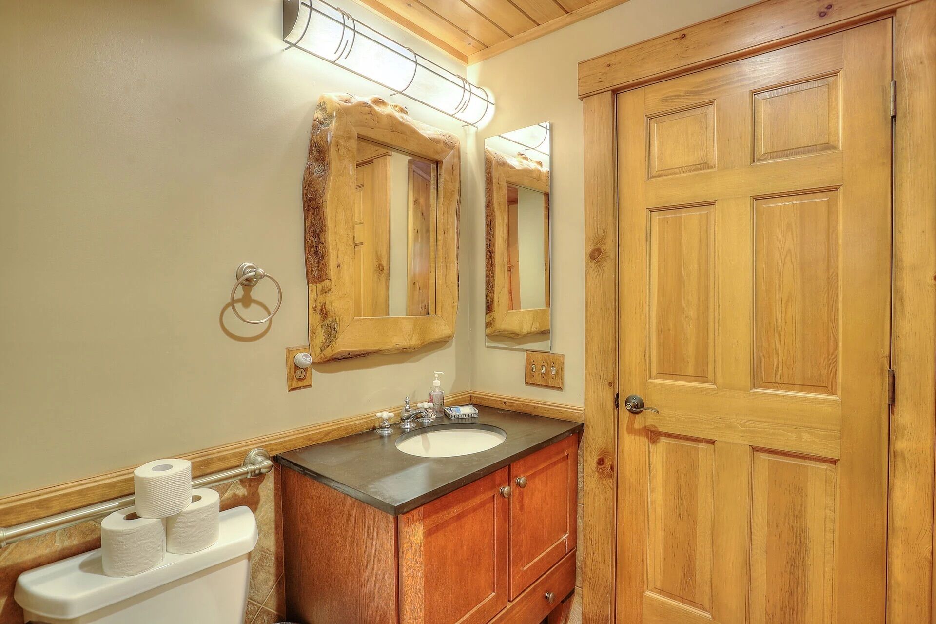 Une salle de bain avec un meuble-lavabo en bois, un évier et un grand miroir. On y trouve des rouleaux de papier toilette accrochés au mur et un porte-serviette.