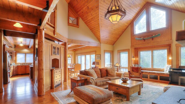 Amplia sala de estar con pisos de madera, un sofá grande, una mesa de centro y una chimenea. Varias ventanas ofrecen luz natural y vistas al exterior.