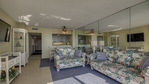 Un salon avec deux canapés à motifs floraux, une table basse et une table à manger avec des chaises. On peut apercevoir une cuisine à travers la porte vitrée, un téléviseur fixé au mur et un ventilateur de plafond.