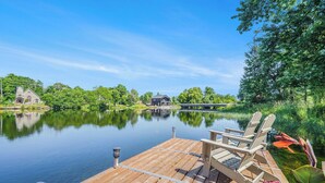 A wooden dock with two Adirondack chairs, a calm lake, and houses in the background.