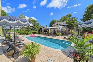 A swimming pool with lounge chairs and umbrellas, surrounded by greenery and a covered patio area.