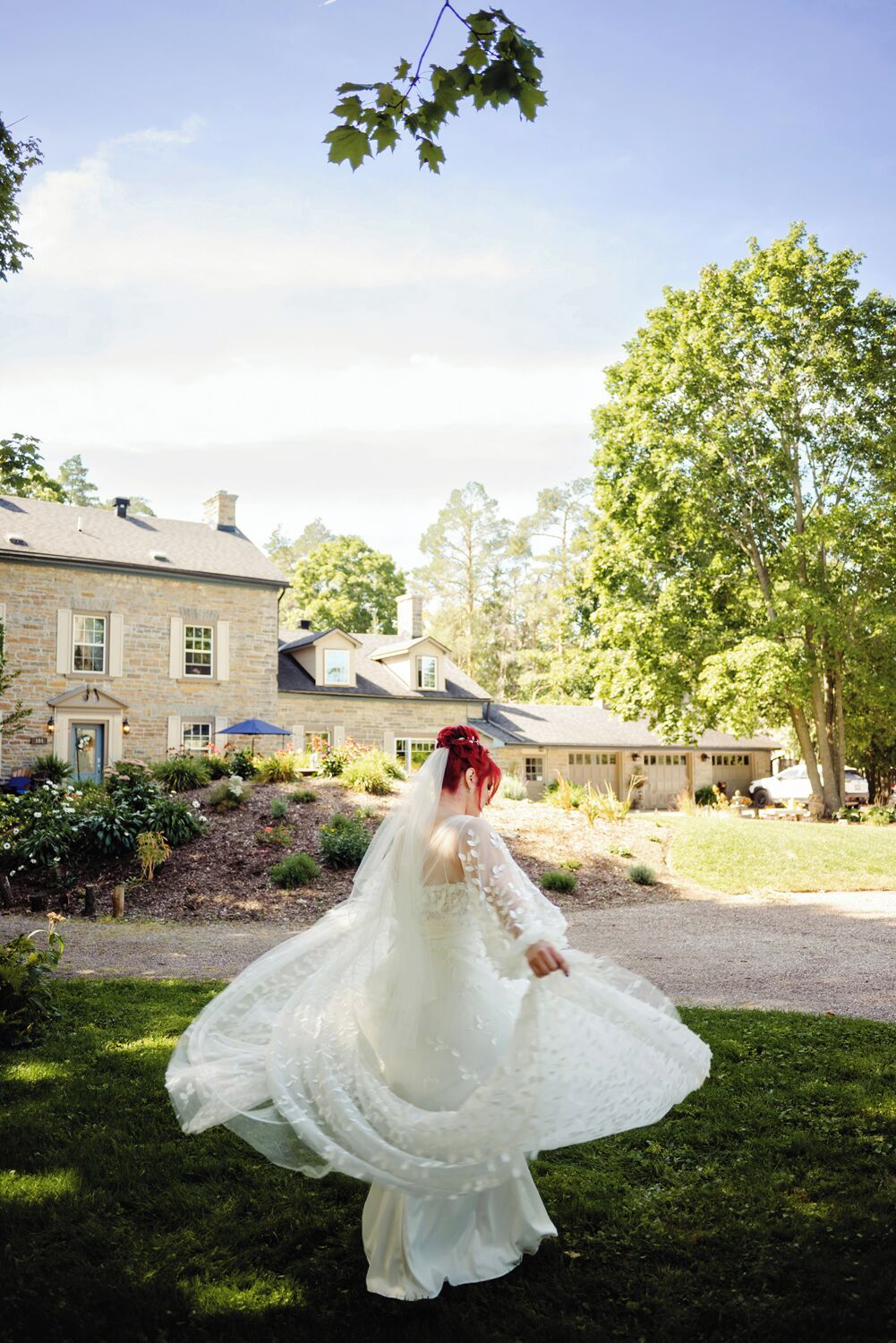 Una novia con un vestido de novia blanco y un velo largo, de pie en un jardín con un edificio de piedra al fondo.