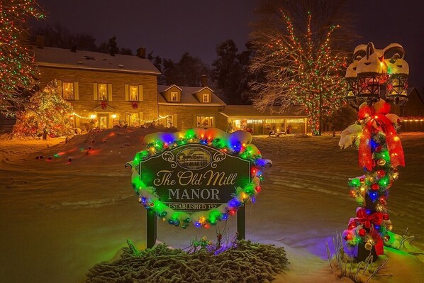 A snow-covered landscape with a sign for "The Old Mill Manor" established in 1852, surrounded by festive Christmas decorations.