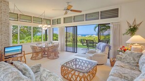 A spacious living room with a television, wicker furniture, a glass coffee table, and a view of the ocean.