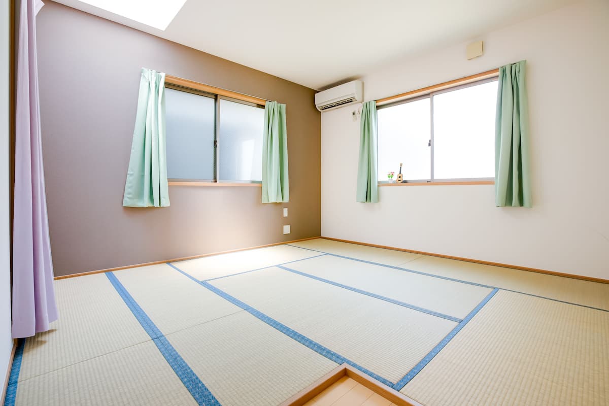 A room with tatami flooring, two windows with green curtains, and an air conditioning unit on the wall.
