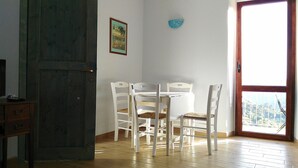 Une pièce avec un plancher en bois, une table blanche avec quatre chaises, une porte donnant sur une vue agréable et un petit tableau encadré accroché au mur.