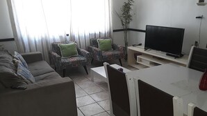 Een woonkamer met een bank, twee fauteuils met een patroon, een televisie en een eettafel.