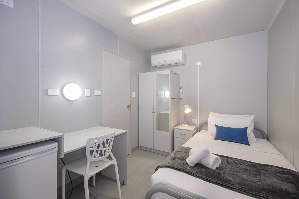 Une chambre d’hôtel moderne équipée d’un bureau, d’une chaise, d’une armoire, d’une table de chevet, d’une lampe, d’un lit et d’un appareil de climatisation.