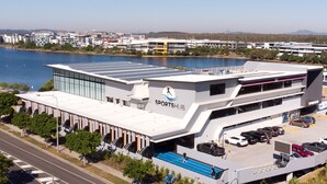 Een modern gebouw met een zonnepaneelinstallatie op het dak, een overdekte parkeerplaats, en een uitzicht op een meer en stadsbeeld op de achtergrond.