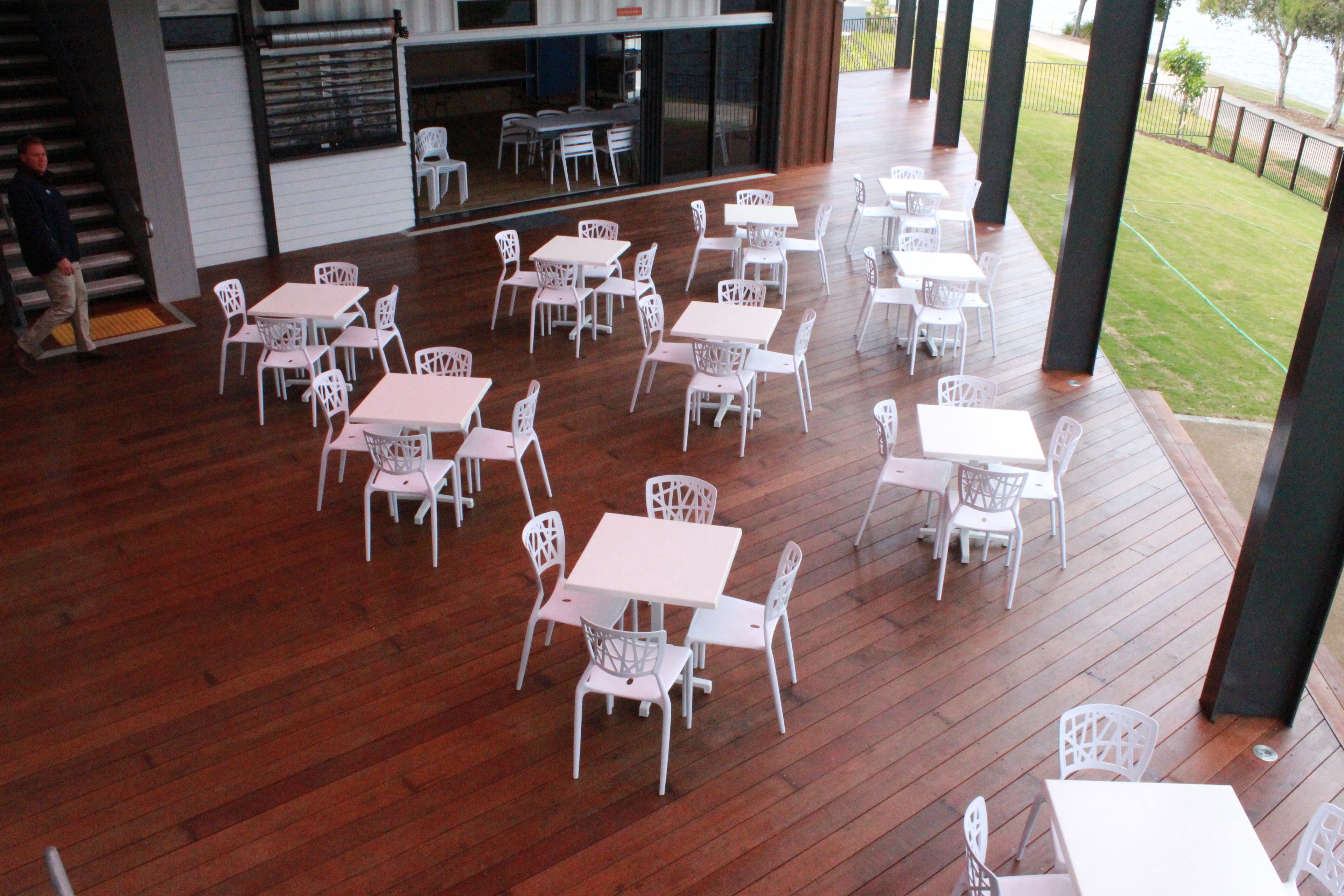 An outdoor patio with multiple white tables and chairs, wooden flooring, and a view of the surrounding area.