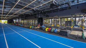 Een ruime fitnessruimte met blauwe vloerbedekking, krachttrainingsapparatuur en een hoge ruimte met zichtbare ventilatiekanalen.