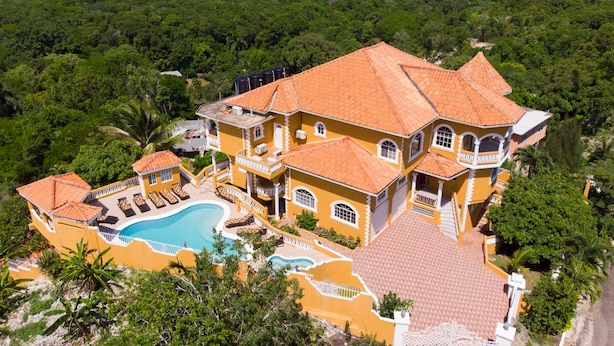 A large, yellow building with a swimming pool and multiple balconies, surrounded by lush greenery and a view of the ocean.