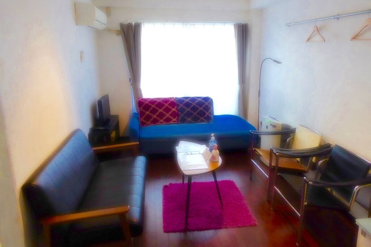 Ein kleines Hotelzimmer mit einer Couch, einem Tisch mit einer Flasche, einem Sessel, einem Schreibtisch mit Lampe und einem Fenster mit Vorhängen.