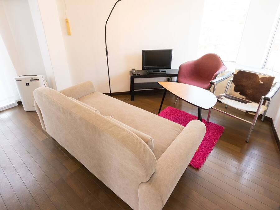 A living room with a beige sofa, a small table, a TV on a stand, a red chair, and a window.
