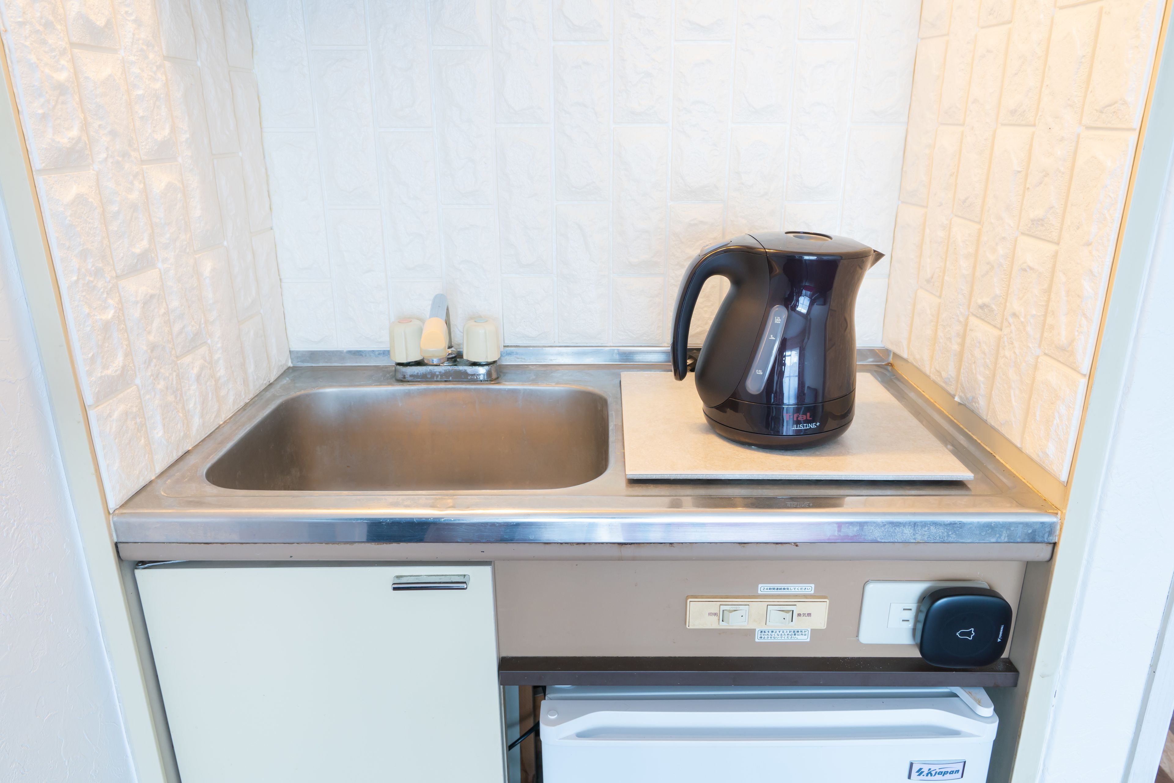 A compact kitchen with a stainless steel sink, a black electric kettle, and a built-in refrigerator.
