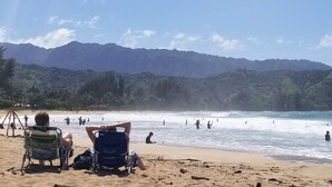 A beach with people in the water and on the sand, mountains in the background, and a clear sky. - Breathtaking Oceanfront Condo (Princeville)