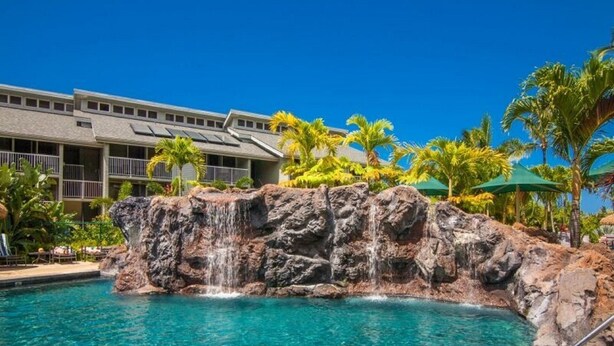 A pool with a waterfall feature, surrounded by palm trees and a building with balconies. - Breathtaking Oceanfront Condo (Princeville)