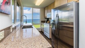 A kitchen with a stainless steel refrigerator, a microwave, and a sink. There is a window with a view of the ocean. - Breathtaking Oceanfront Condo (Princeville)