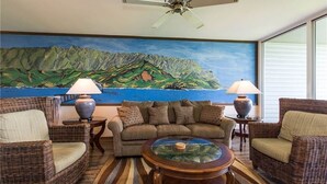A living room with wicker furniture, a glass coffee table, and a painting of a coastal landscape. - Breathtaking Oceanfront Condo (Princeville)