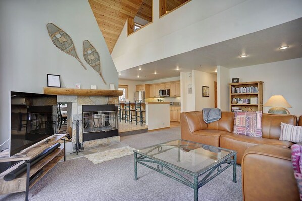 A spacious living area with a fireplace, a glass coffee table, a leather sofa, and a kitchen in the background.