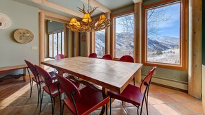 Een eetgedeelte met een grote houten tafel en rode stoelen, een kroonluchter en ramen met uitzicht op de bergen.