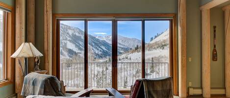 A cozy room with a large window offering a mountain view, a wooden floor, and a lamp on a side table.