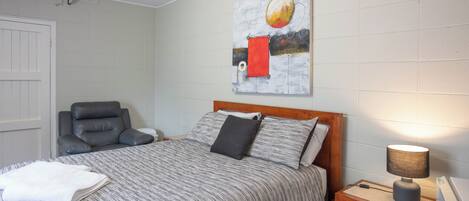 A neatly made bed with a striped bedspread, a wooden nightstand with a lamp, a gray leather chair, and a wall-mounted painting.