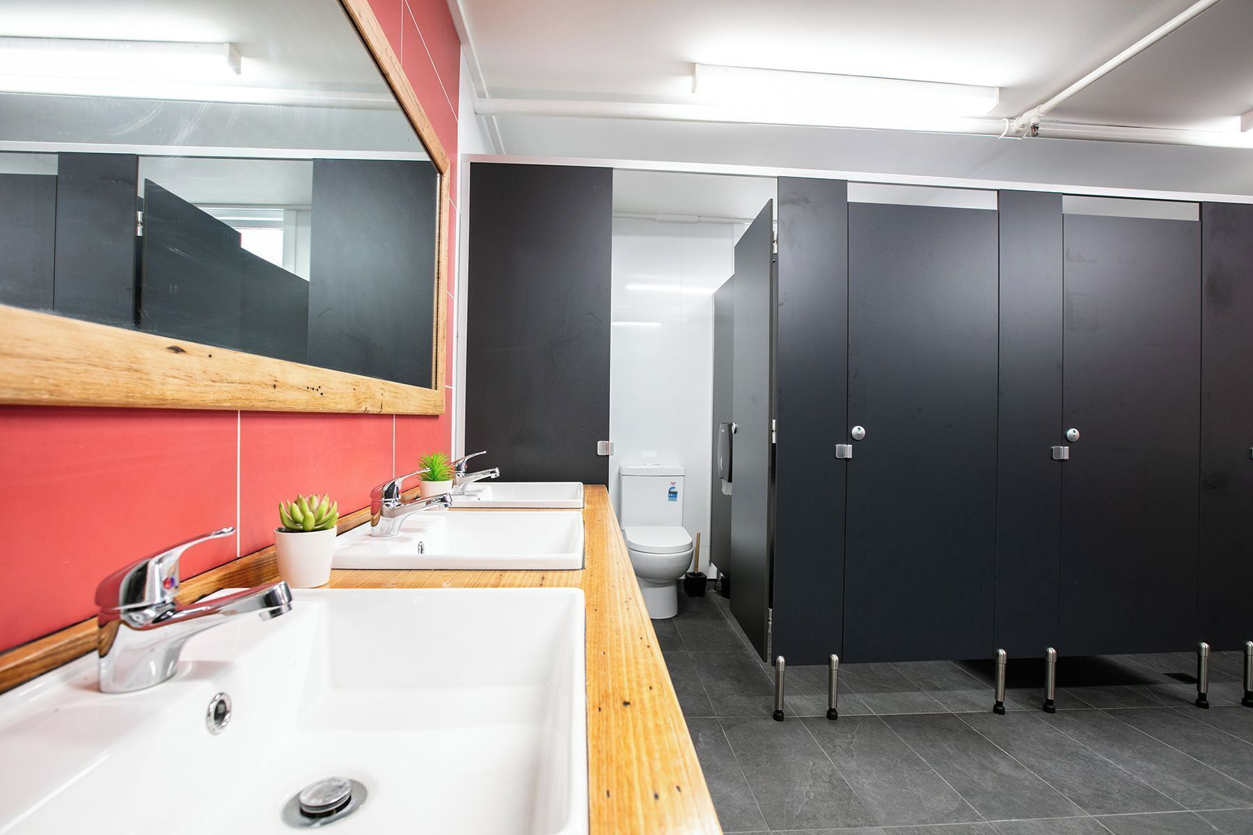Un baño moderno con un lavamanos blanco, un espejo y un inodoro. Hay cabinas negras y una pared roja.