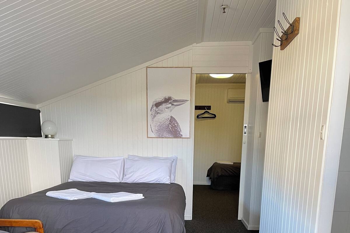 Ein Schlafzimmer mit einem Bett, einem Holzsessel, einem Kleiderhaken und einem gerahmten Bild an der Wand.