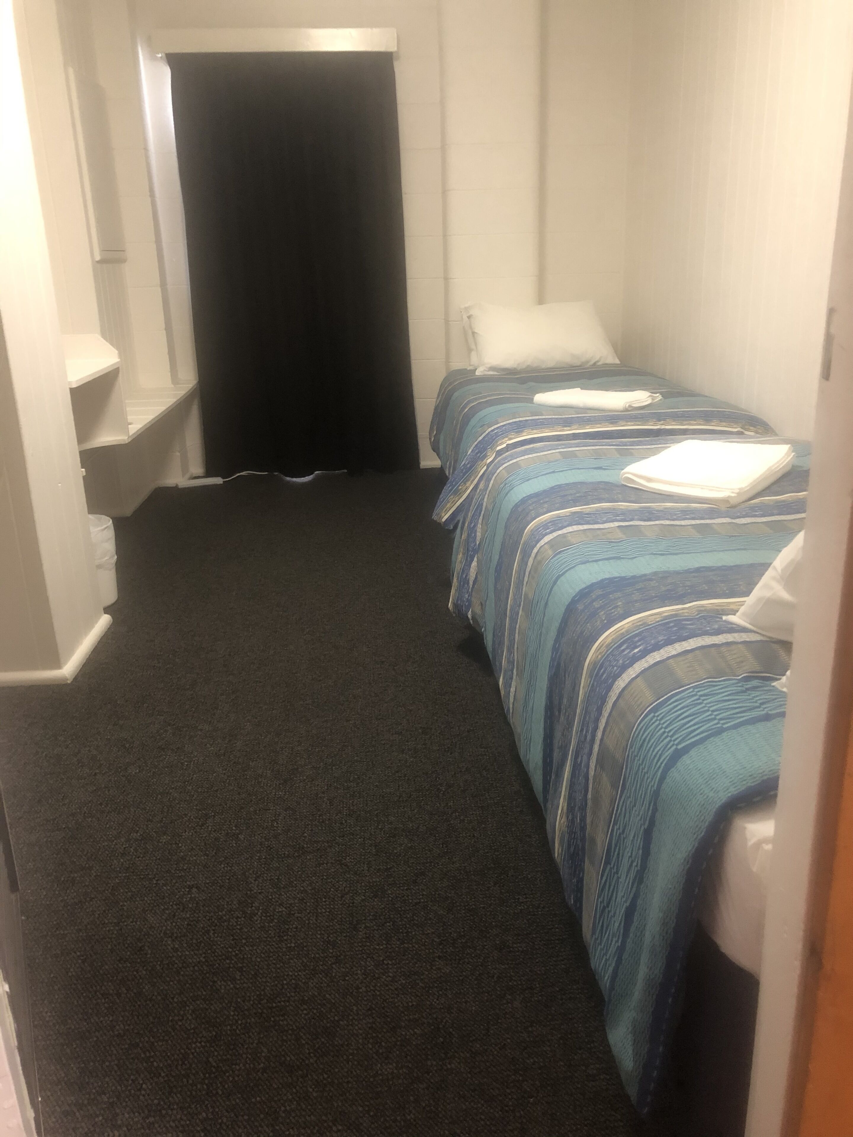 A small, single-bed room with a dark carpet, a striped bedspread, and a white pillow.
