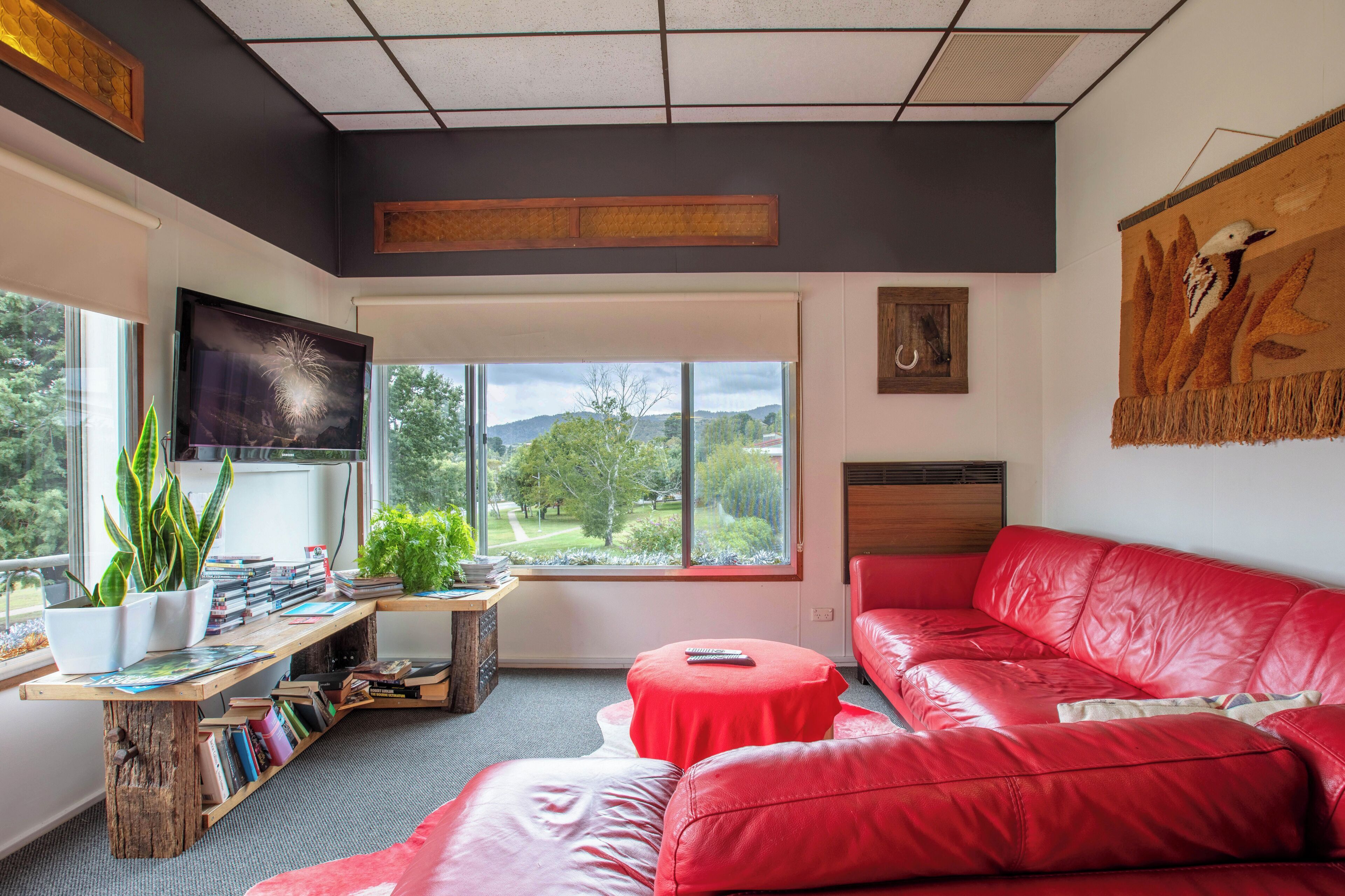 A modern living room with a red leather sofa, a wooden coffee table, and a bookshelf filled with books. There is a large window offering a view of the outdoors.
