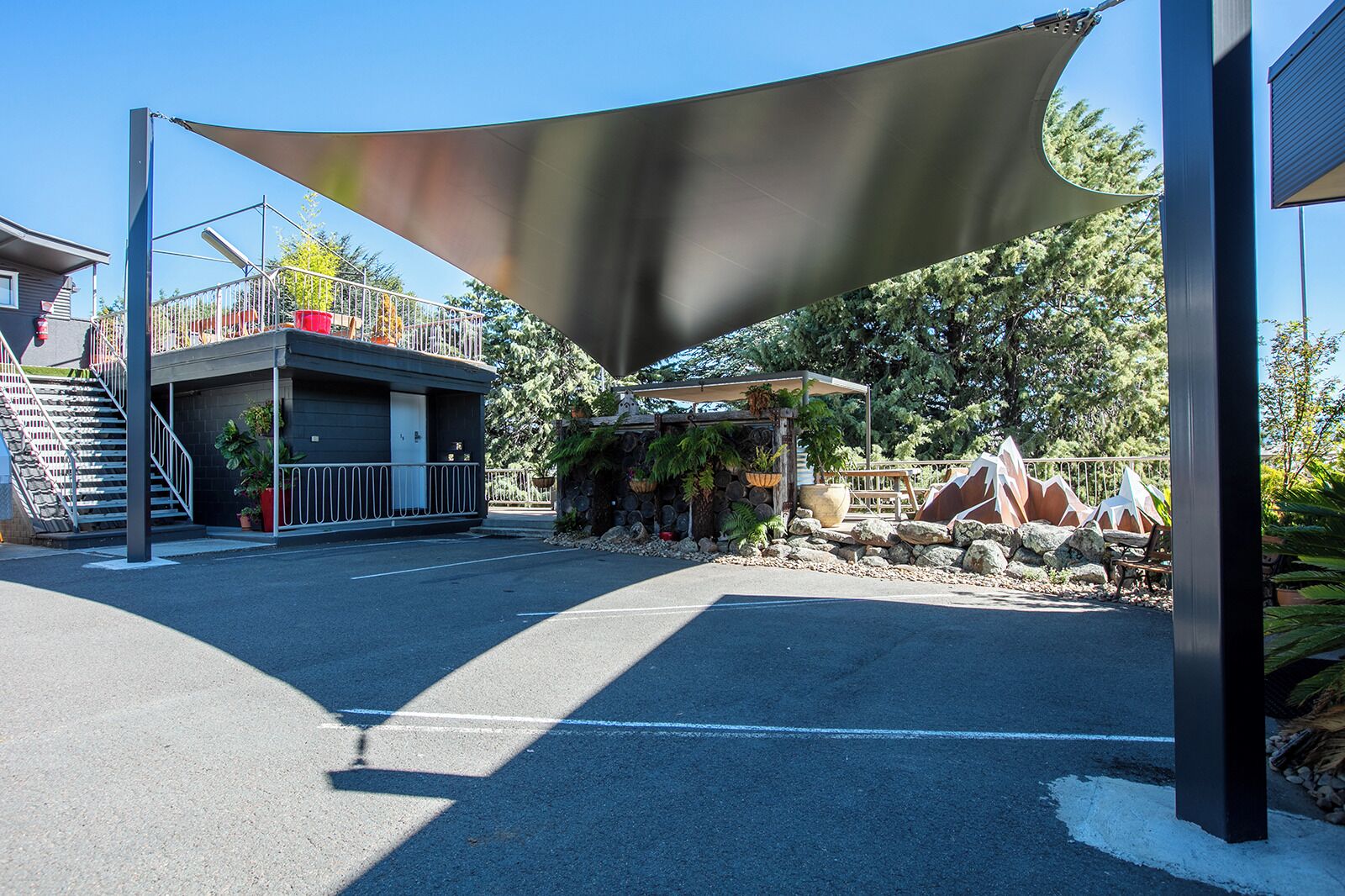 A parking area with a canopy structure, a building with a balcony, and a stone wall with plants.