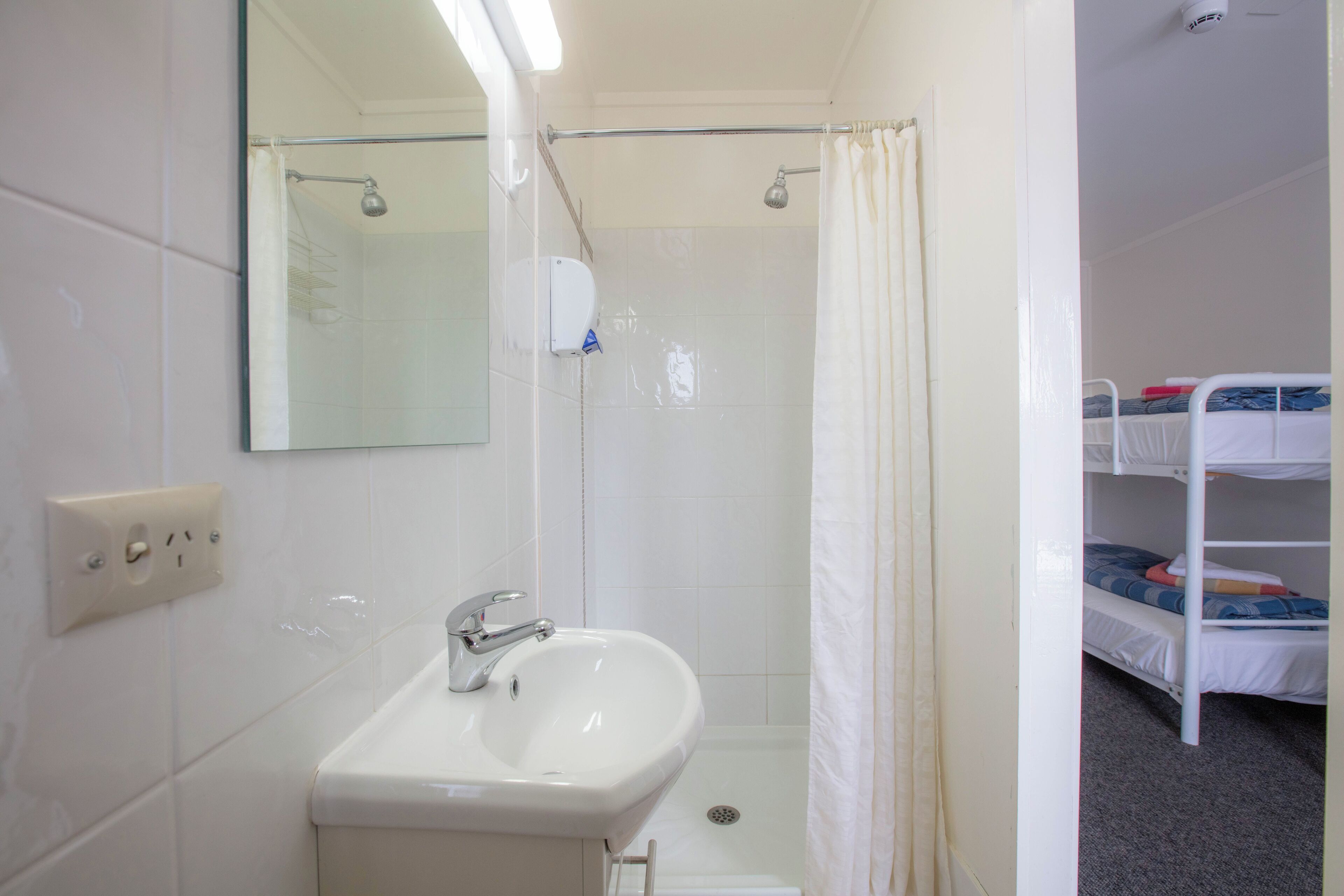 A bathroom with a sink, mirror, shower, and a bunk bed in the adjacent room.