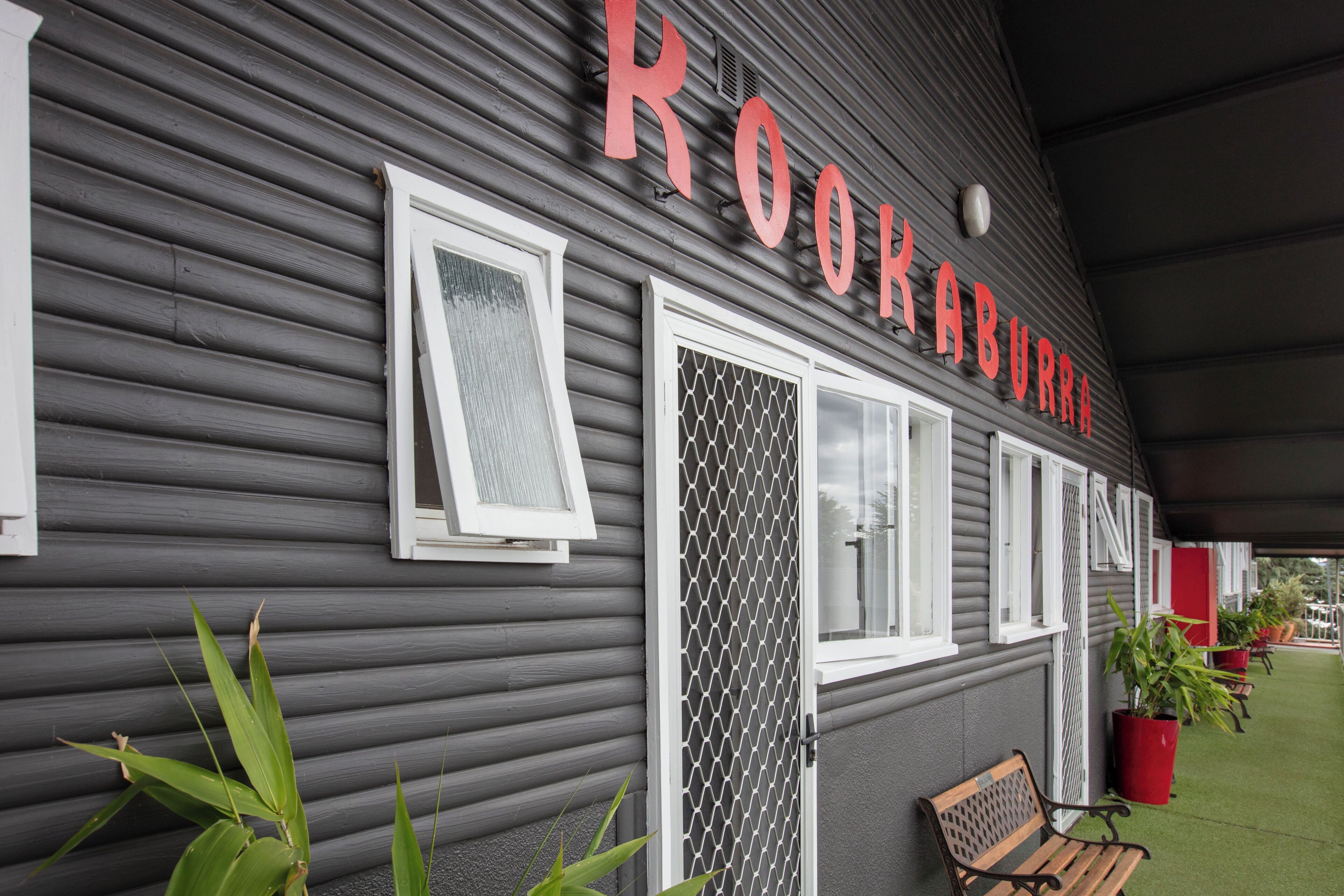 A building with the name "ROOKABURRA" in large letters. There are two windows with white frames, one with a mesh screen and one with a solid door. There is a bench and potted plants outside.