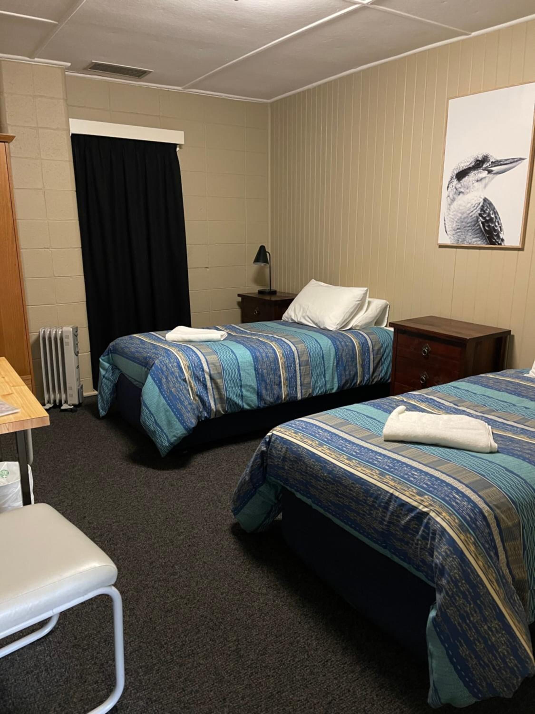 Une chambre d’hôtel avec deux lits, un bureau, une chaise, un radiateur, une table de chevet et un tableau au mur.