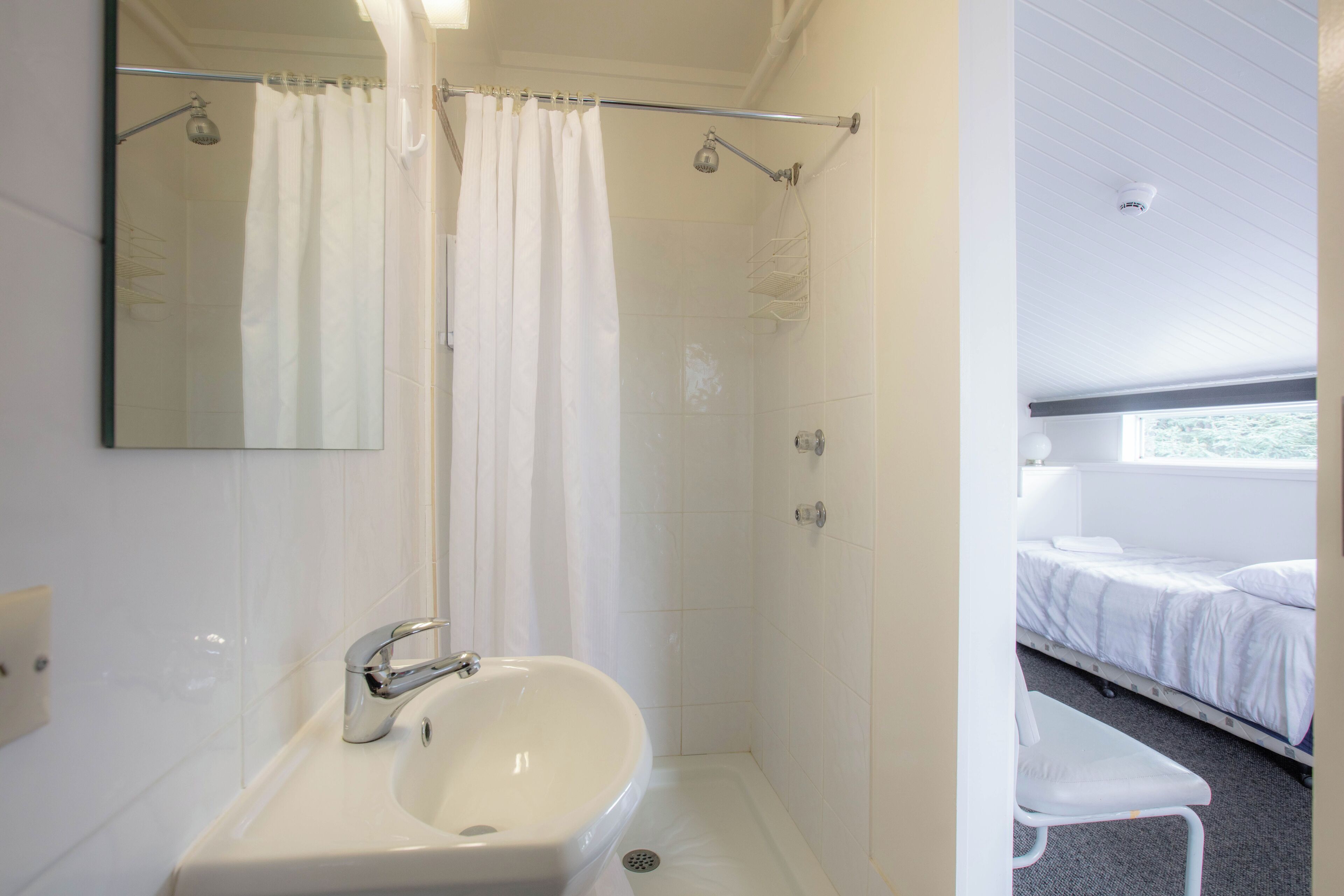 Ein Badezimmer mit Dusche, Waschbecken und einem Bett, das durch eine geöffnete Tür zu sehen ist.