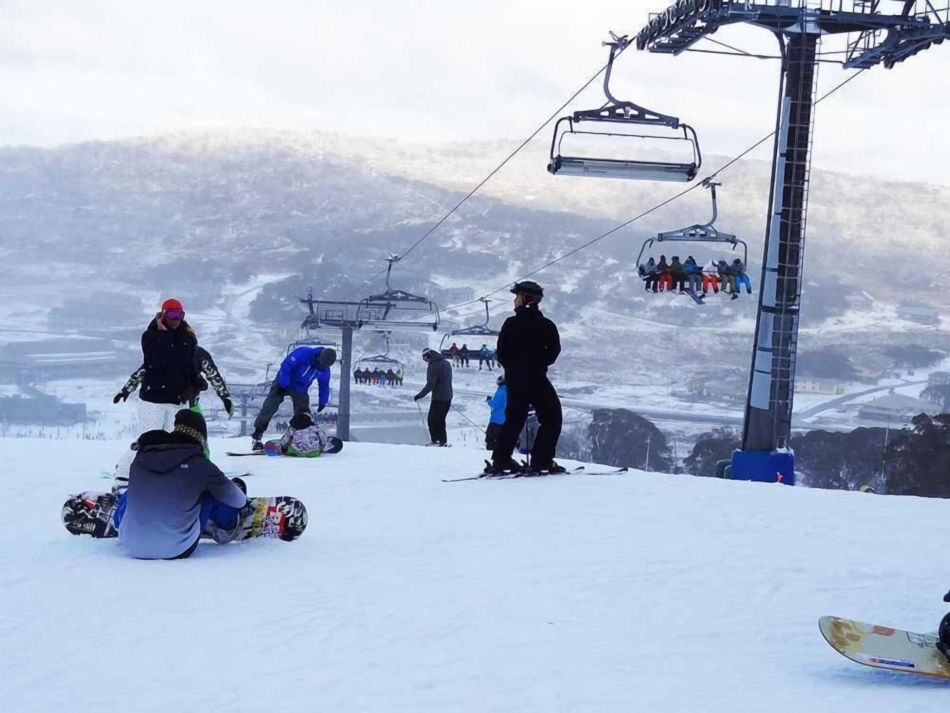 A ski lift with people waiting and a snowboarder sitting on the ground.