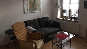Un salón con un sofá gris oscuro, un sillón de mimbre, una mesa de centro de vidrio con un mantel individual rojo, una lámpara colgante y una ventana con vistas a edificios y una calle.