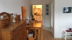 Un comedor con una mesa y sillas de madera, una cocina al fondo y una pequeña mesa blanca con un mantel individual rojo.