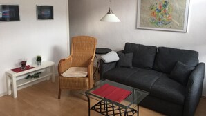 Una sala de estar con un sofá gris oscuro, una silla de mimbre, una mesa de centro de vidrio con un paño rojo y una mesita auxiliar blanca, además de un cuadro en la pared.