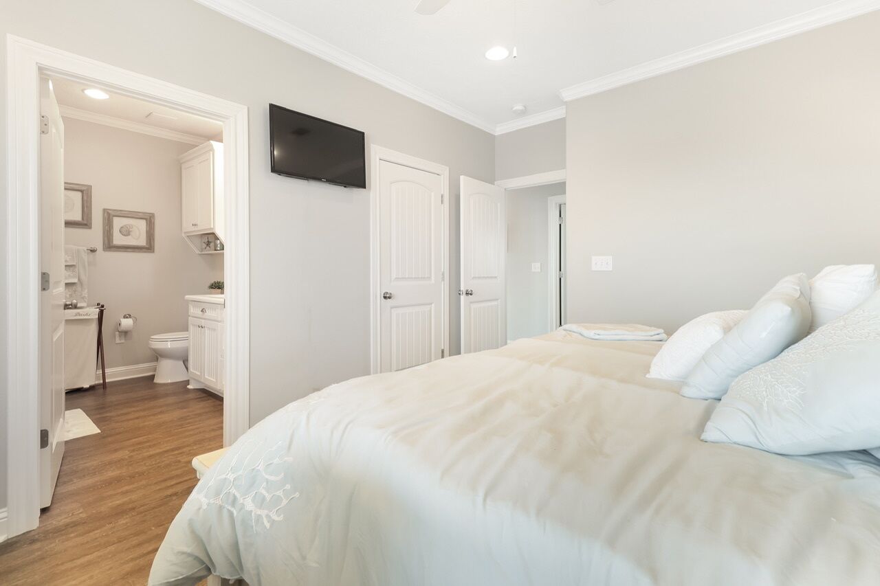 A bedroom with a large bed, white walls, and a flat-screen TV mounted above the door.