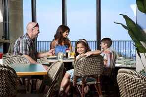 Une famille est assise à une table, près d’une fenêtre donnant sur l’océan, et prend son repas.