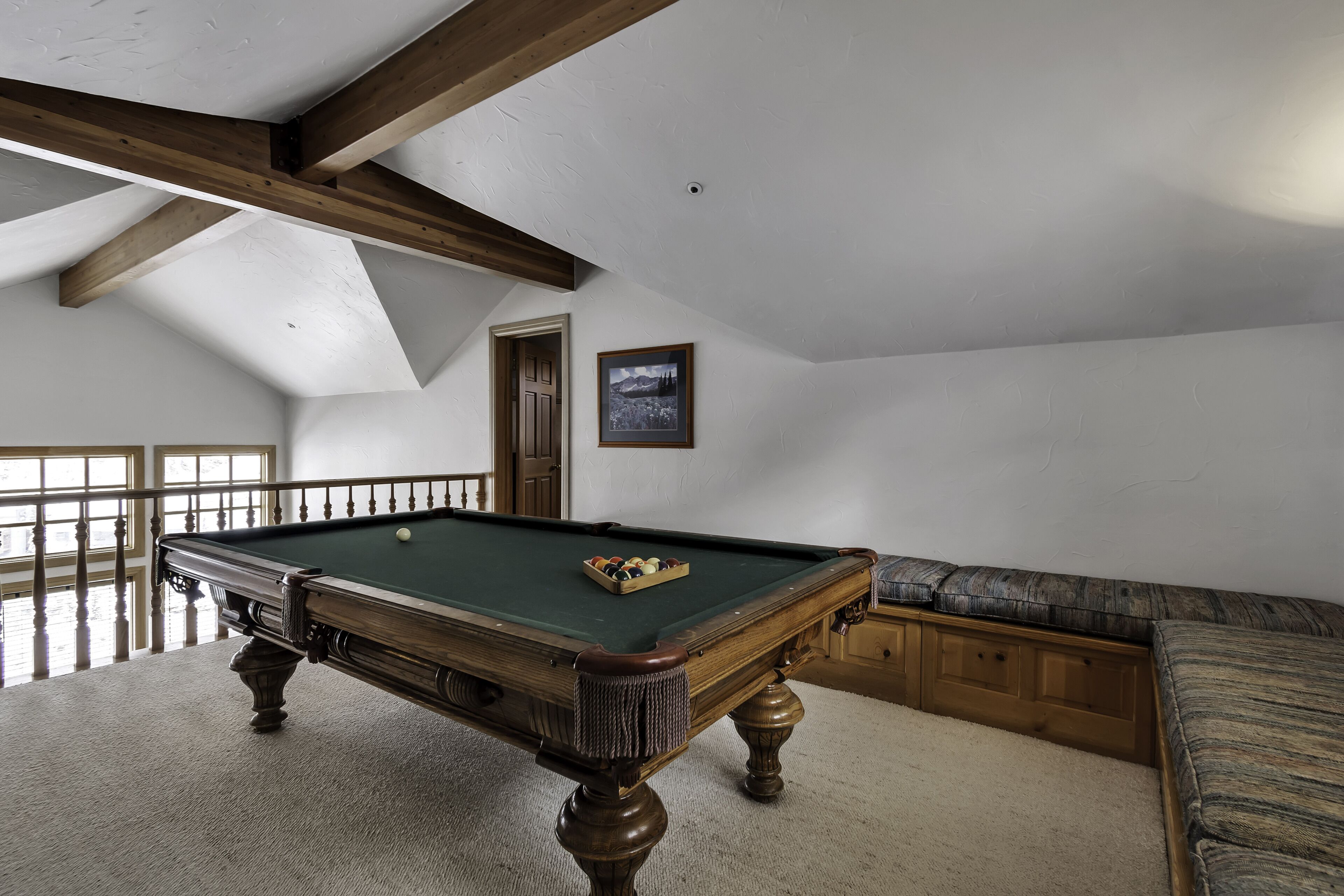 A pool table in a room with a sloped ceiling, wooden beams, a cushioned bench, and a door.